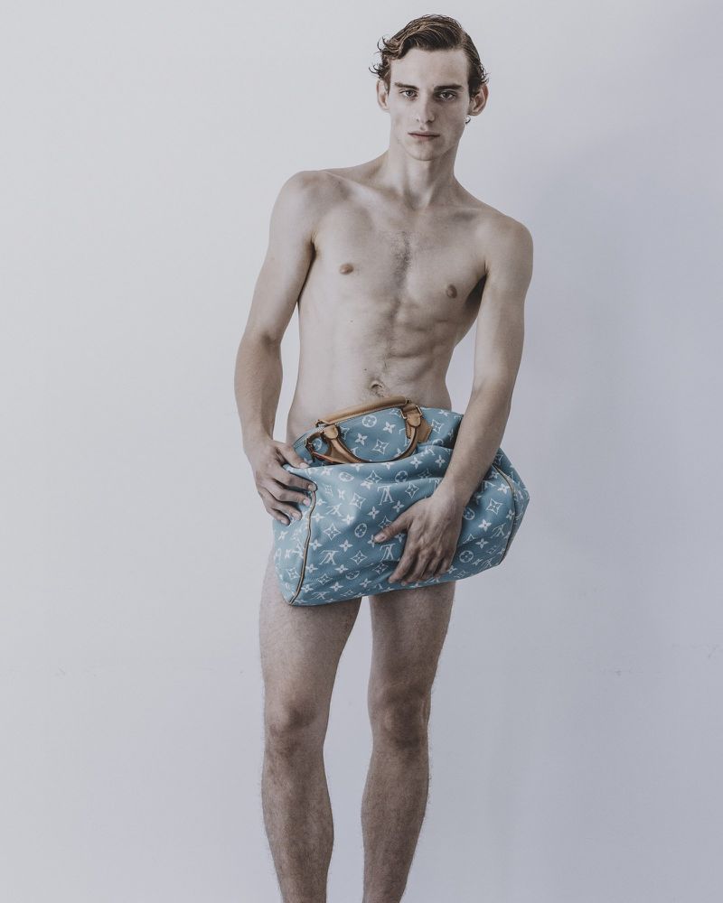 Shirtless male model holding a light blue monogram travel bag against a plain grey studio backdrop