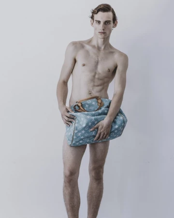Shirtless male model holding a light blue monogram travel bag against a plain grey studio backdrop