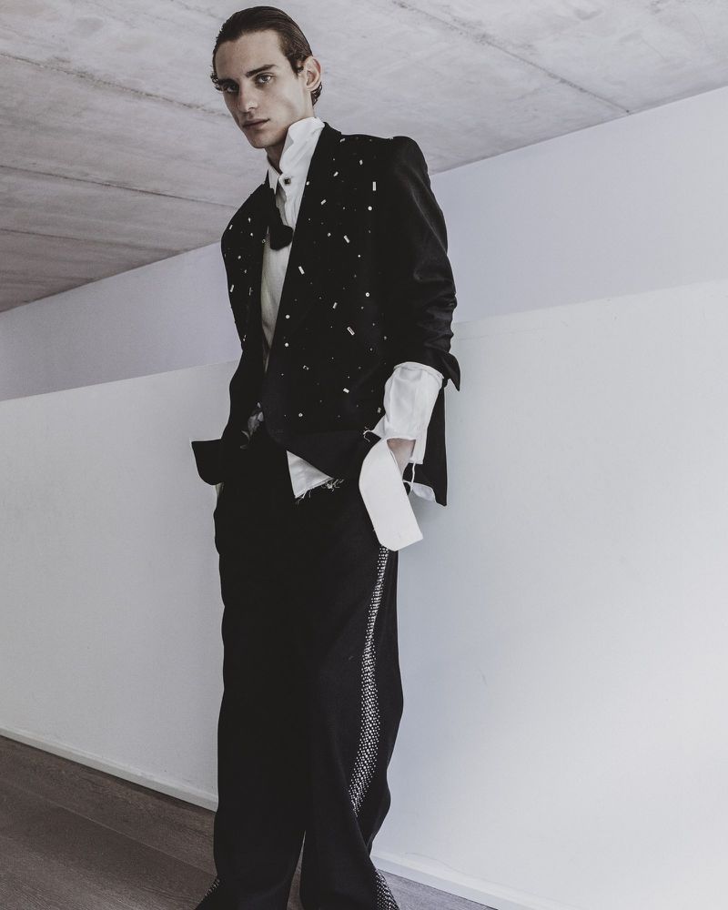 Full-length portrait of a male model wearing a black embellished blazer, white shirt, and black trousers against a concrete wall.