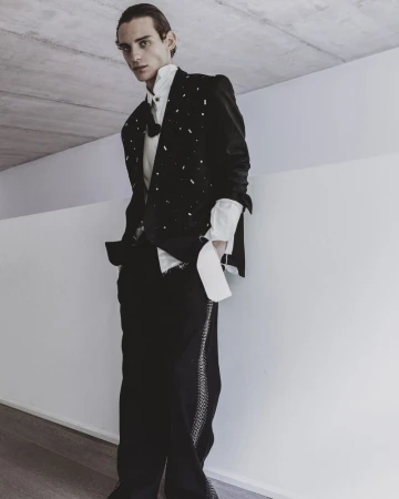 Full-length portrait of a male model wearing a black embellished blazer, white shirt, and black trousers against a concrete wall.