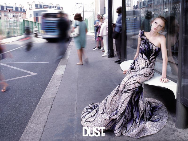Model in a beaded purple and black evening gown seated on a bench along a busy city street.