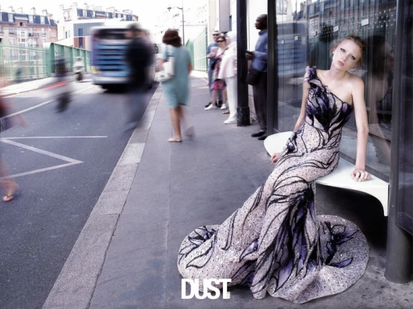 Model in a beaded purple and black evening gown seated on a bench along a busy city street.