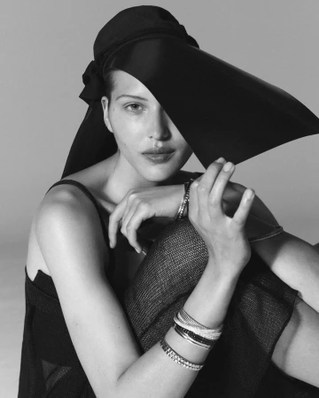 Black-and-white portrait of a woman wearing a large hat, adorned with bracelets, sitting in a photo studio with a neutral background.