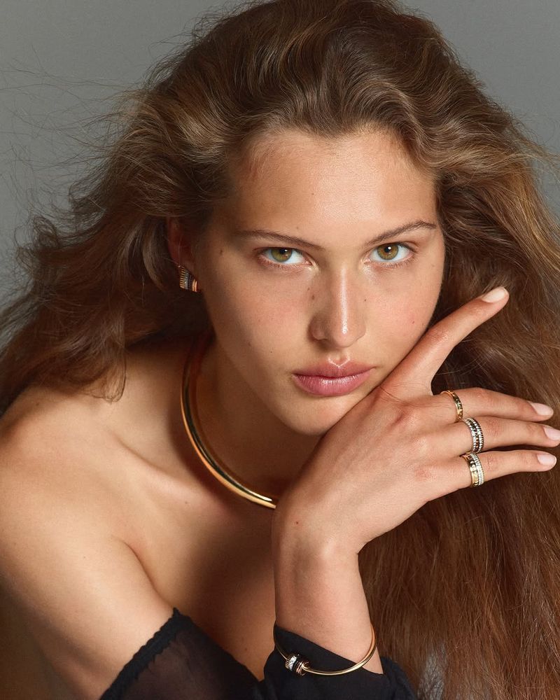 Close-up studio shot of a young adult woman with long brown hair wearing gold jewelry on her neck, wrist, and fingers against a neutral background.