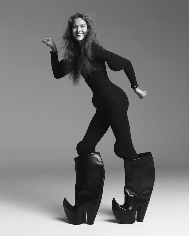 Black and white studio portrait of a smiling woman in a catsuit and oversized boots, striking a dynamic pose against a plain background.