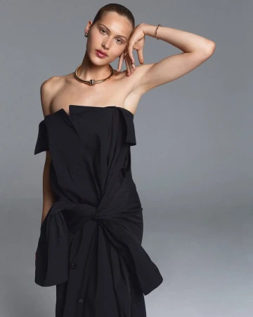 Model Angelina Kendall wearing a black strapless tied dress and gold collar necklace against a grey studio backdrop.