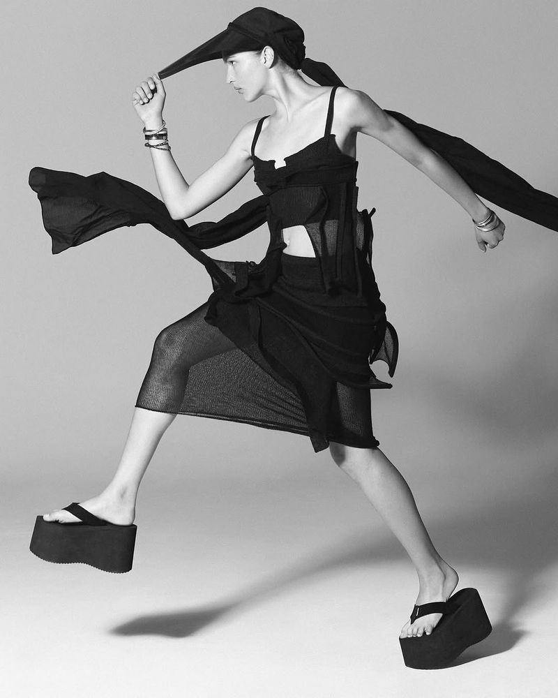 Fashion model in a black outfit, including a headscarf and platform sandals, jumps in a studio setting in a dynamic pose.