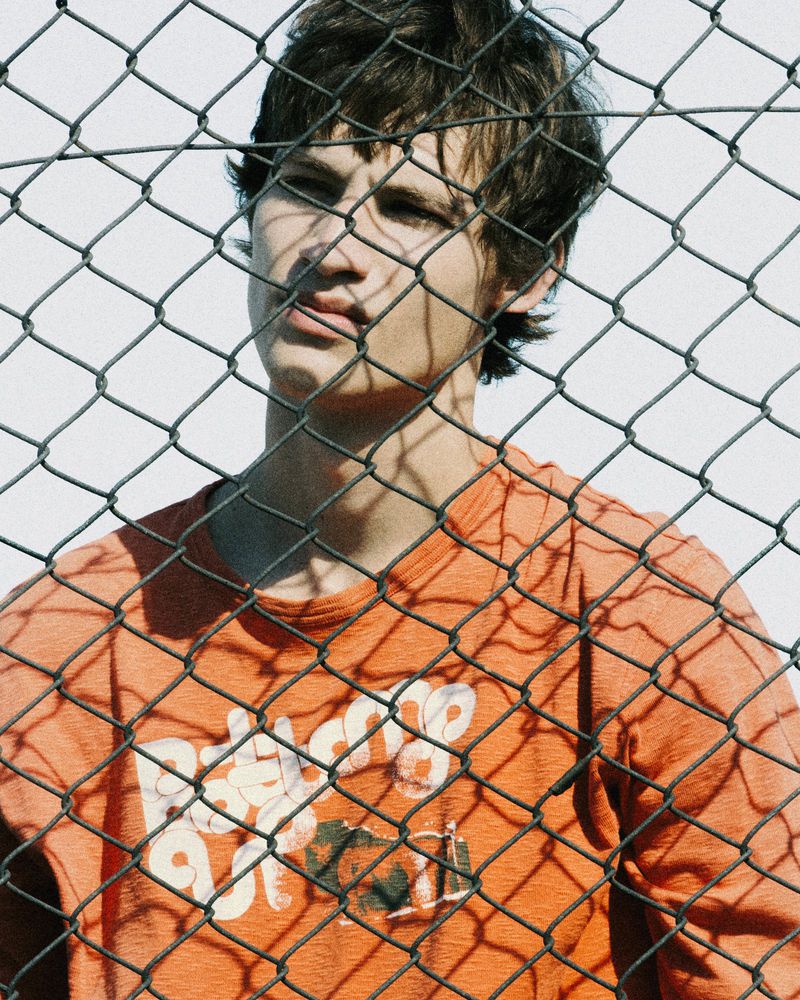Young man wearing an orange t-shirt behind a chain-link fence for Fucking Young editorial