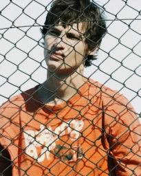Young man wearing an orange t-shirt behind a chain-link fence for Fucking Young editorial