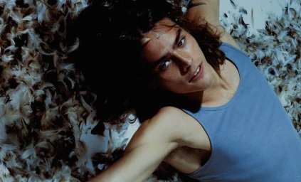 Young male model with long dark hair lying among scattered white and brown feathers wearing a blue ribbed tank top