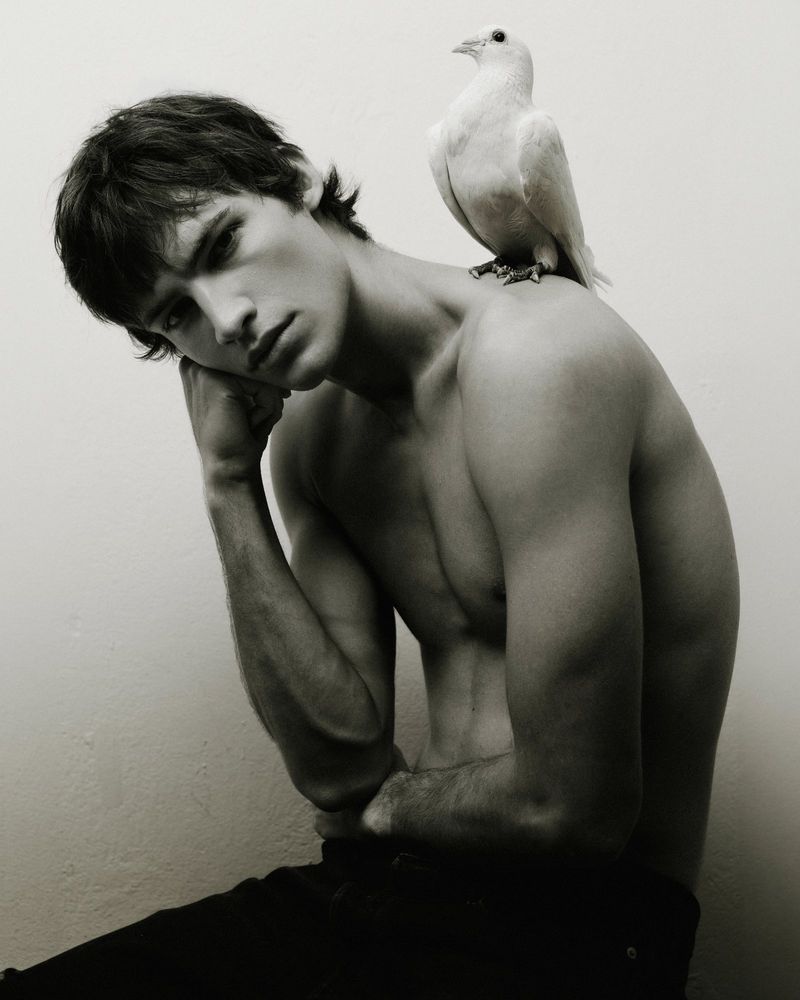 Shirtless young man sitting with a white dove perched on his shoulder against a plain white background