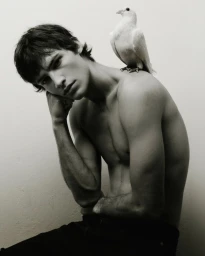 Shirtless young man sitting with a white dove perched on his shoulder against a plain white background
