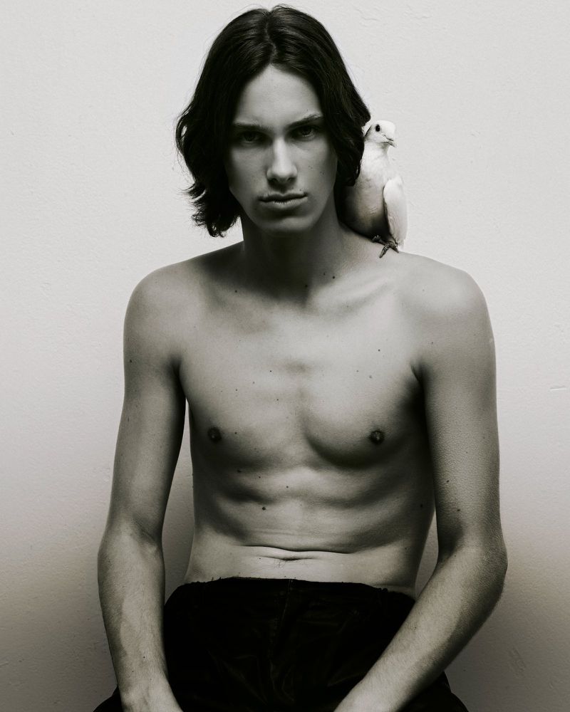 Shirtless young man with dark medium-length hair posing with a white bird perched on his shoulder against a plain white background