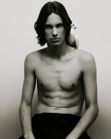 Shirtless young man with dark medium-length hair posing with a white bird perched on his shoulder against a plain white background
