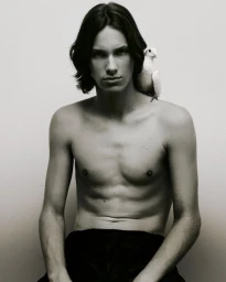 Shirtless young man with dark medium-length hair posing with a white bird perched on his shoulder against a plain white background