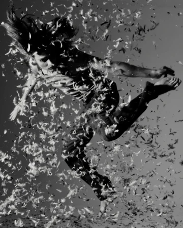 Monochrome action shot of a male model leaping amidst a swirling cloud of falling feathers in a studio setting.