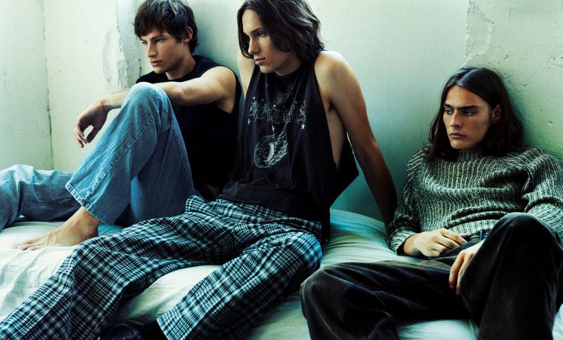 Three male models seated together on a white mattress against a pale green wall in a casual youth fashion editorial.