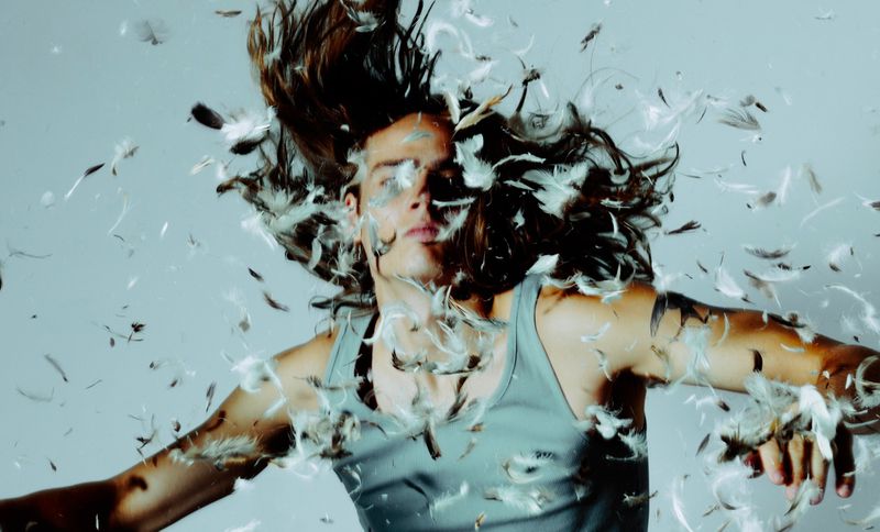 Long-haired man wearing a grey tank top surrounded by floating feathers against a grey studio background