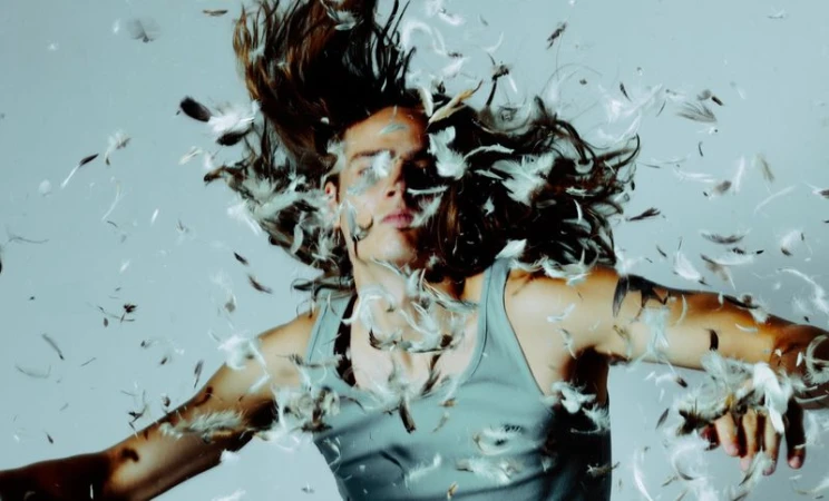 Long-haired man wearing a grey tank top surrounded by floating feathers against a grey studio background