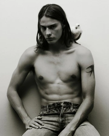Shirtless male model with long dark hair wearing denim jeans with a small white bird perched on his shoulder.