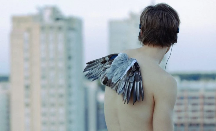 Shirtless young man with small bird wings attached to his back looking out at blurred urban apartment buildings