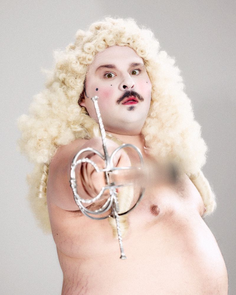 Shirtless man wearing a large blonde curly wig and theatrical makeup pointing a rapier toward the camera.