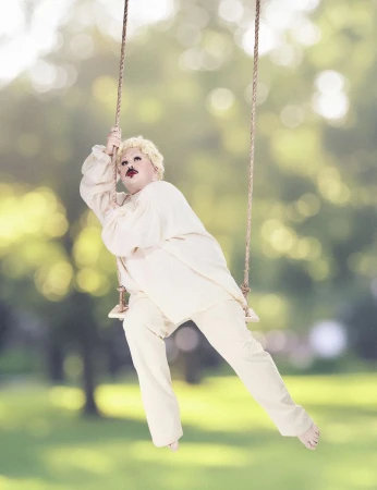 Person in cream-colored clothing with a small mustache riding a rope swing outdoors against a green park background.
