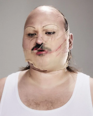 Close-up portrait of a bald man wearing a nude-colored stocking stretched over his face, creating distorted features against a neutral grey studio background.