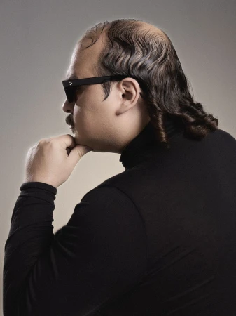 Profile portrait of a man wearing black sunglasses and a black turtleneck resting his chin on his hand against a neutral grey background