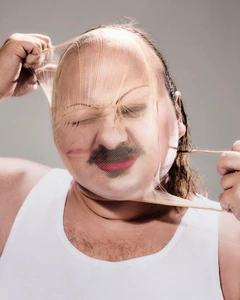 Man wearing a white tank top pulling a nude-colored stocking tight over his face in a studio portrait.