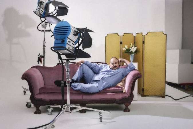 Man reclining on a vintage pink velvet sofa in a brightly lit white studio with professional lighting equipment