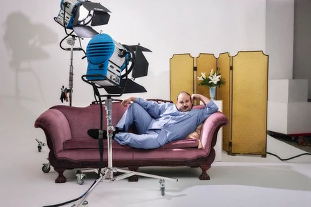 Man reclining on a vintage pink velvet sofa in a brightly lit white studio with professional lighting equipment