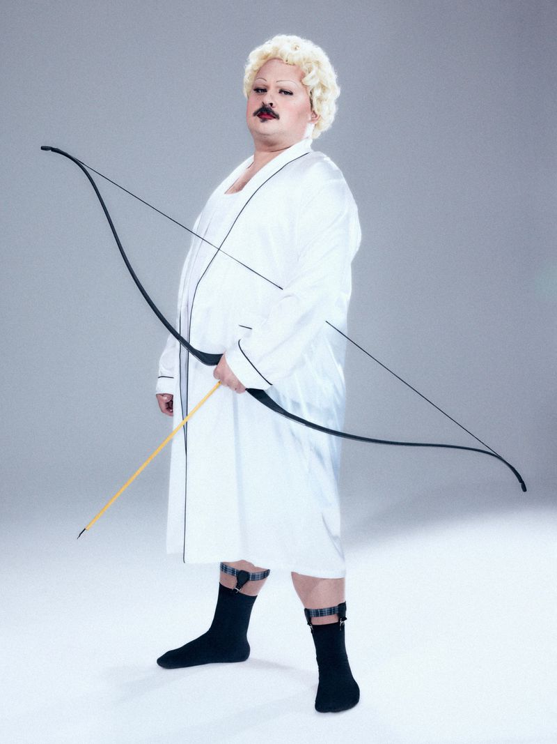 Person wearing white robe and blonde curly wig holding a large bow against a grey seamless studio background.