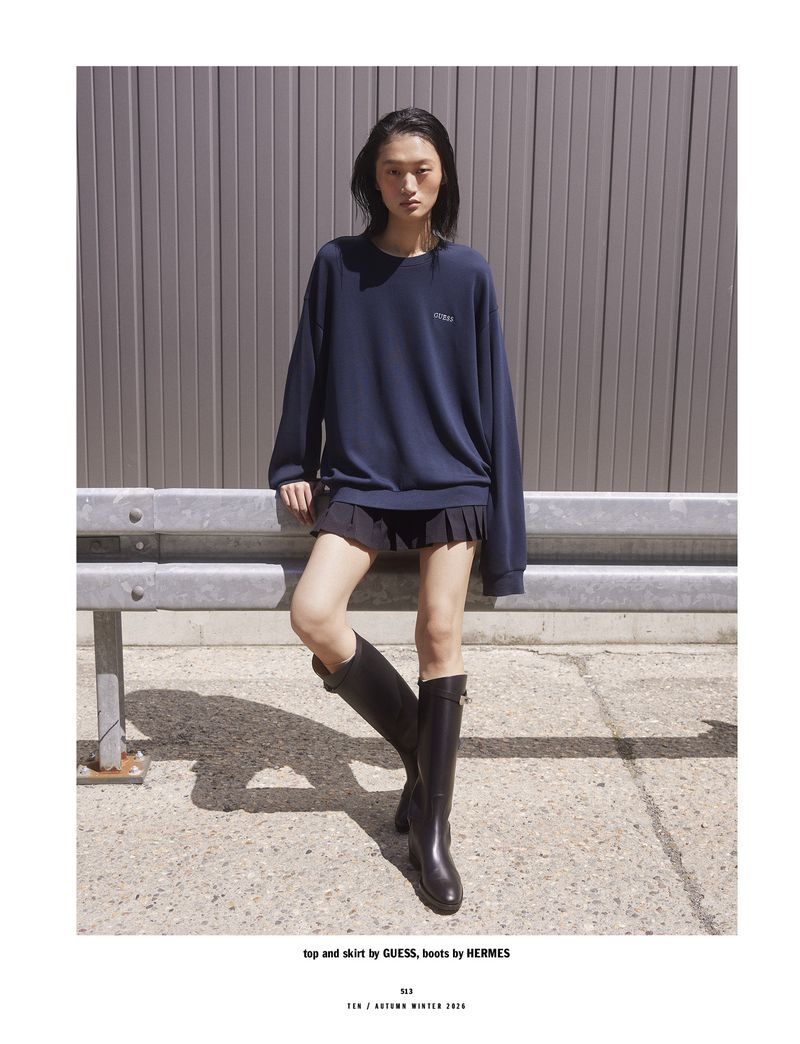 Model Yilan Hua leaning against a metal guardrail wearing a navy blue sweatshirt and black pleated miniskirt with tall leather boots.