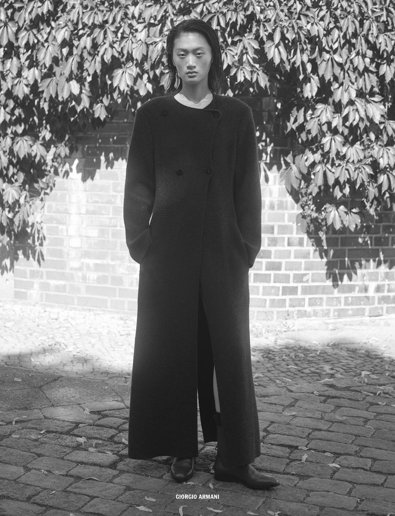 Model Yilan Hua wearing a long black coat standing outdoors on cobblestones in 10 Magazine editorial Day Shift by Mark Pillai.