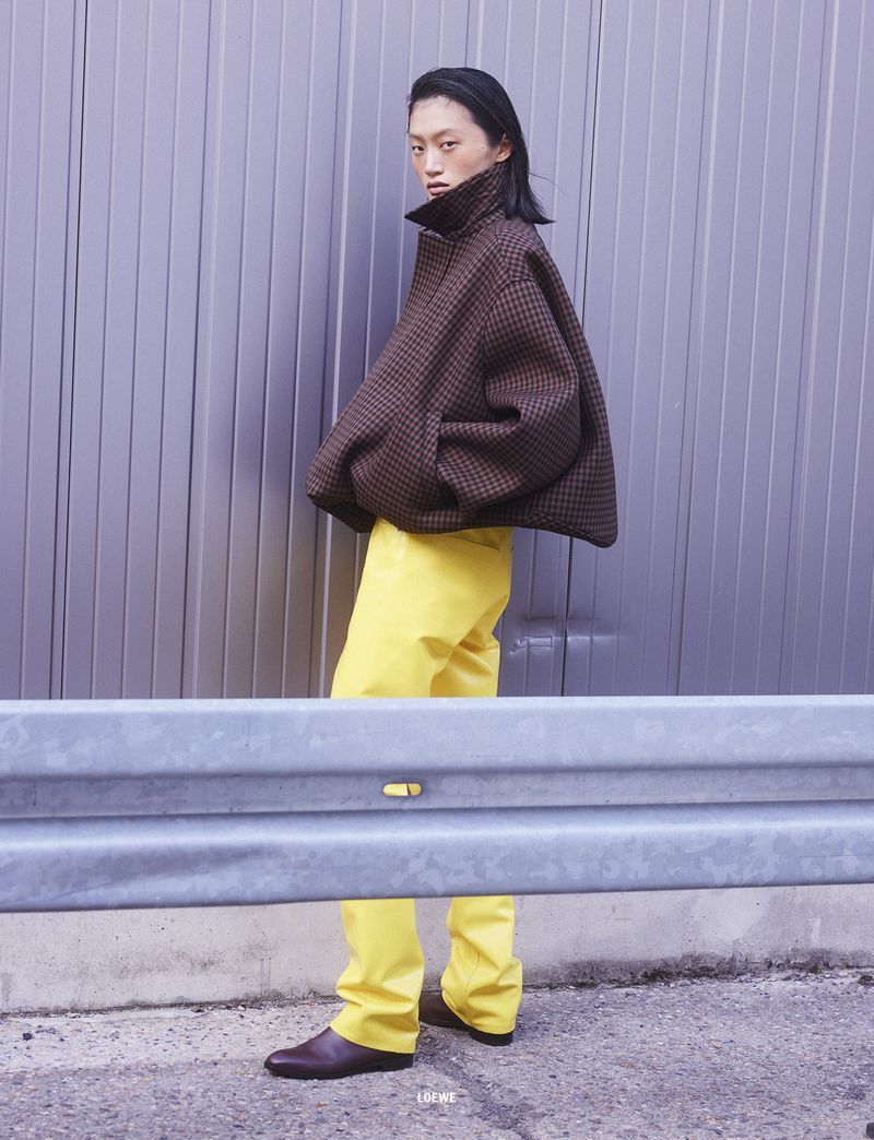 Model Yilan Hua wearing a brown textured jacket and yellow trousers against a corrugated metal wall