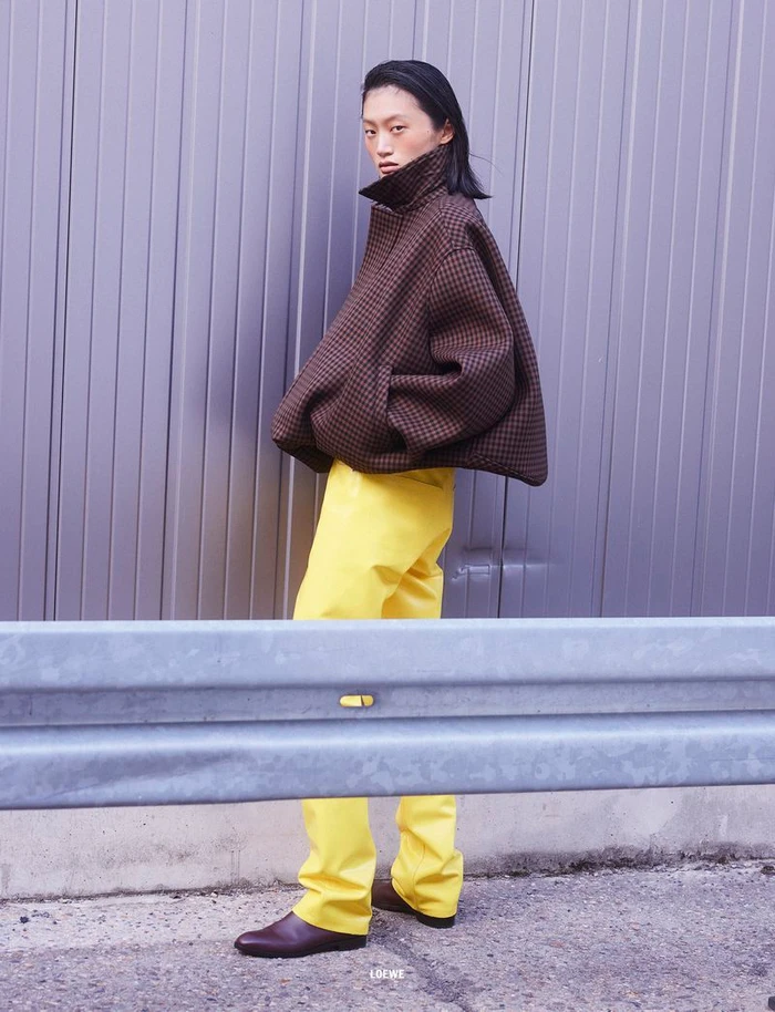 Model Yilan Hua wearing a brown textured jacket and yellow trousers against a corrugated metal wall