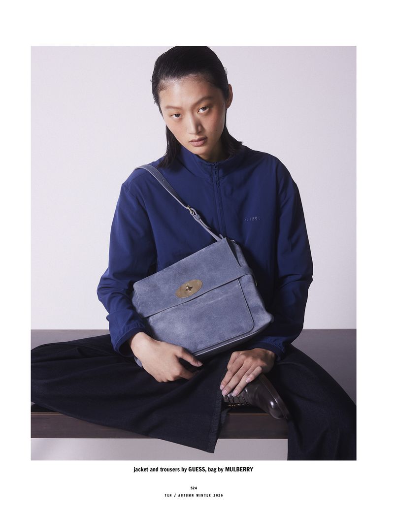 Model Yilan Hua wearing a blue jacket and holding a crossbody bag against a simple studio backdrop.