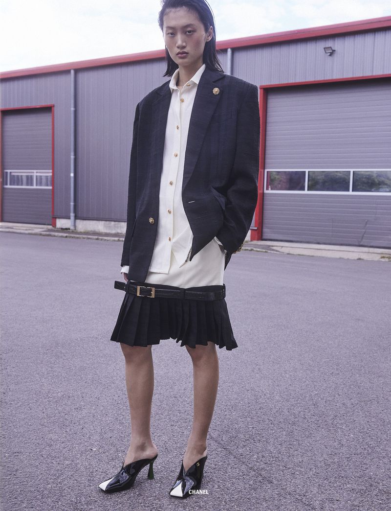Model wearing an oversized black blazer and pleated skirt standing on an asphalt street in front of a corrugated warehouse building