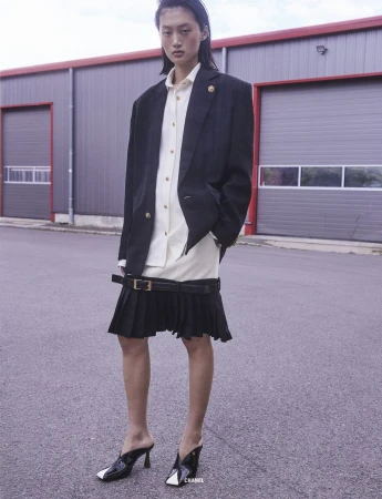 Model wearing an oversized black blazer and pleated skirt standing on an asphalt street in front of a corrugated warehouse building