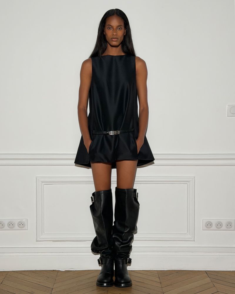 Model standing against a white wall wearing a black sleeveless dress and tall black leather boots