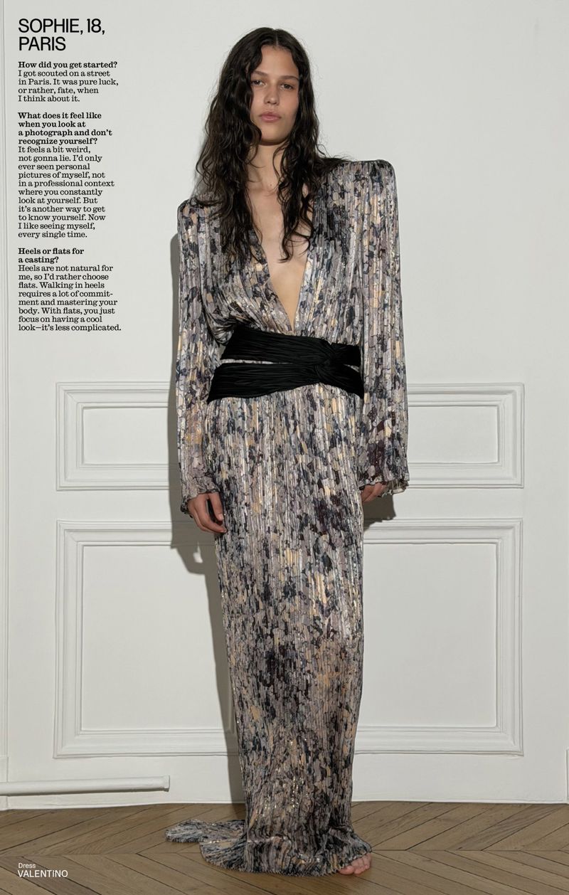 Model in a metallic printed gown with a black sash standing against white wall molding