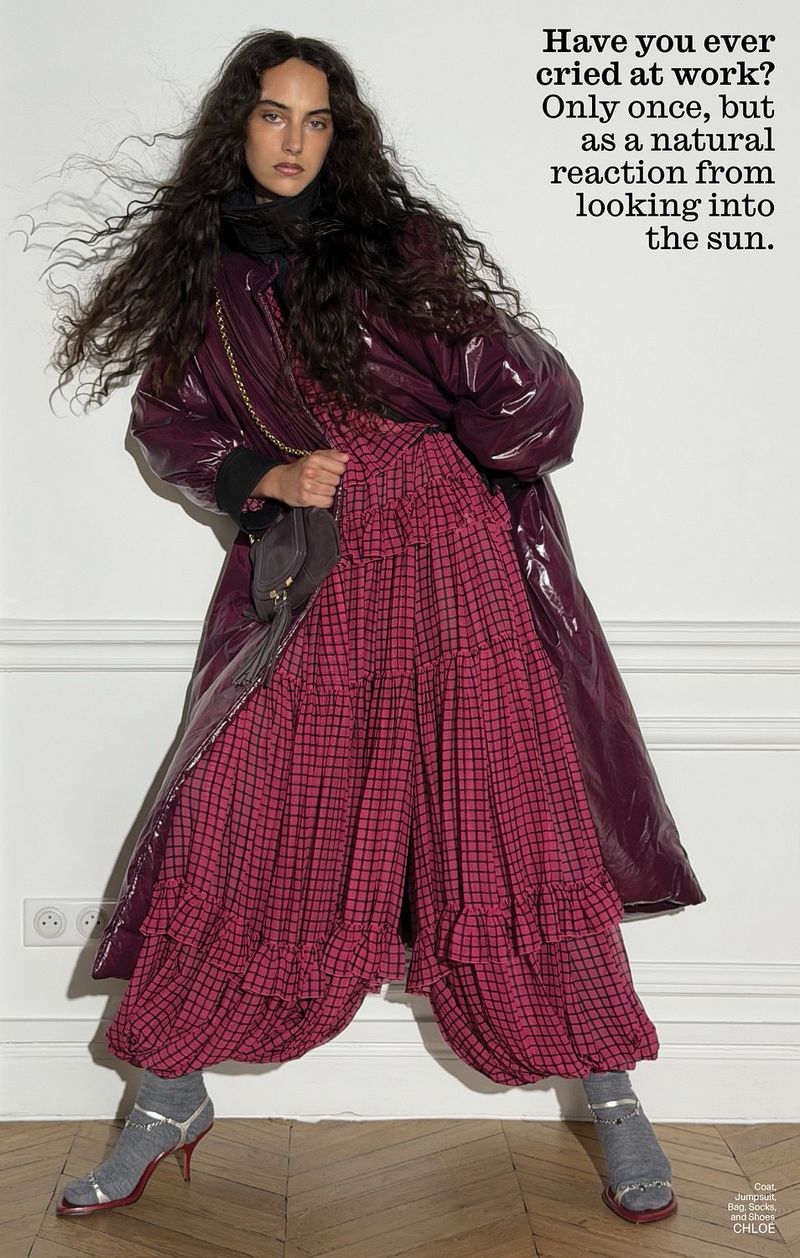 Model with long dark curly hair wearing a glossy purple coat over a pink patterned dress and strappy heels against a white wall.