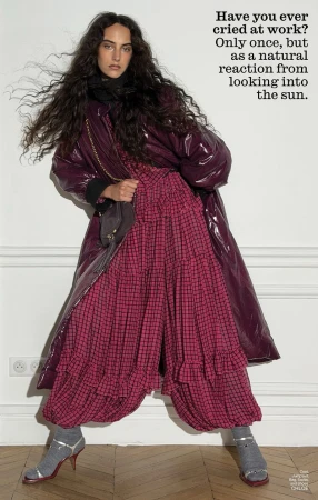 Model with long dark curly hair wearing a glossy purple coat over a pink patterned dress and strappy heels against a white wall.