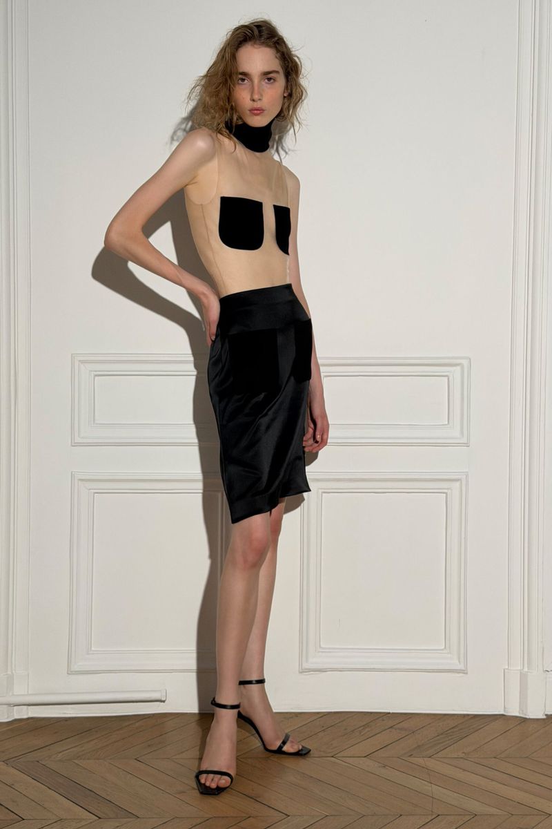 Model posing against a white paneled wall wearing a sheer top with black pockets and a black skirt