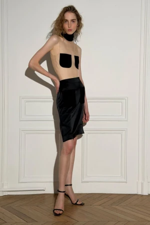 Model posing against a white paneled wall wearing a sheer top with black pockets and a black skirt
