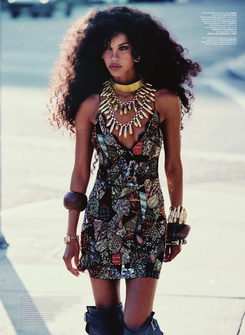 Model Daiane Sodré with voluminous dark curly hair wearing a patterned dress and layered gold necklaces outdoors.