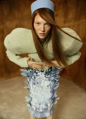 Model with long red hair wearing a light blue hat, structured green jacket, and textured blue patterned skirt against textured background