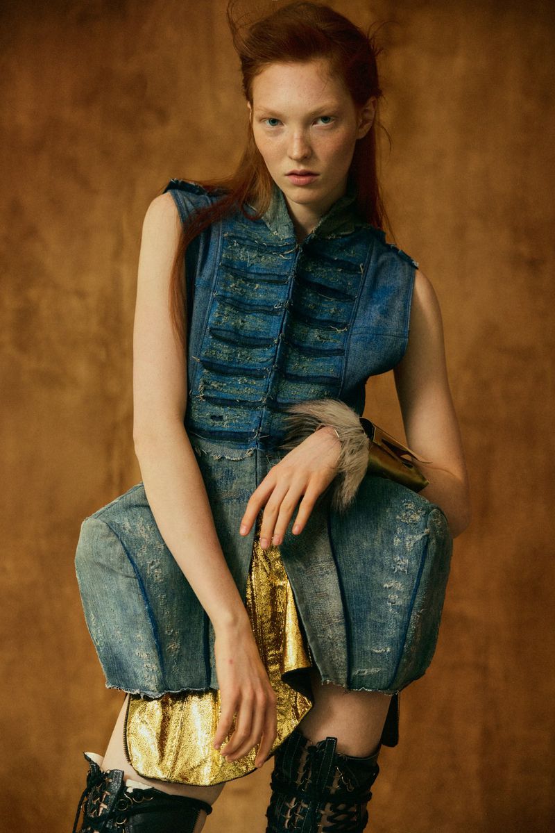 Model with red hair wearing a structured blue denim vest and matching trousers against a textured golden brown background.