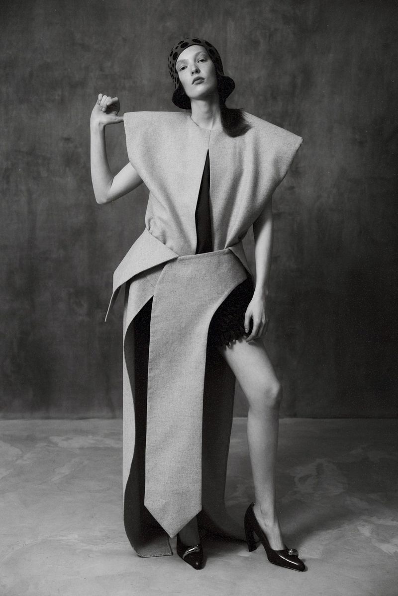 Model in structured avant-garde coat wearing a patterned head covering and black heels against a mottled background